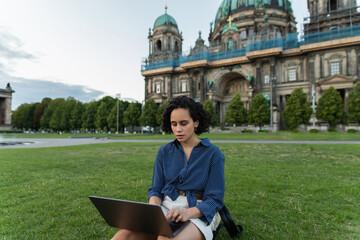 young woman using laptop and sitting on grass near blurred cathedral in berlin during trip. © LIGHTFIELD STUDIOS