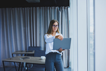A young smiling business woman is standing in the office by the window with a laptop in her hands. Woman manager in glasses and white shirt