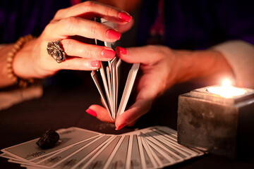 Fortune teller female hands shuffling a deck of tarot cards, during a reading. Close-up with candle light, moody atmosphere. - obrazy, fototapety, plakaty