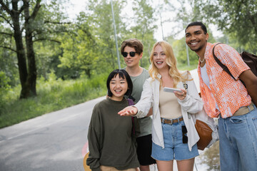 smiling woman with smartphone pointing with hand near interracial friends on road.