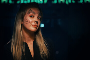 Blonde woman with cat makeup. Charming woman looks cunningly into the camera. Halloween party at a nightclub. All Saints' Night. Garland lights on the background. Photo card with copy space.