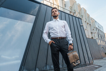 Shot of professional executive man with bag posing around city buildings in daytime.