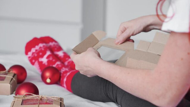 Women's hands packs a gift box, prepare a surprise for the holiday. The girl makes a nice New Year's gift on the bed. The best Christmas present ever. Red toy balls, wrapping paper and Christmas hat.