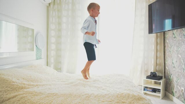 Caucasian baby boy jumping on parent's big bed indoor home in bedroom, looking at empty TV screen with copy space. Fun time at leisure activity. Happy childhood in motion. Cheerful happy moment life.