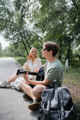 happy blonde woman applauding to boyfriend sitting on road and playing acoustic guitar.