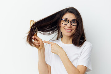 beautiful happy woman in glasses and a white t-shirt combing her long, healthy hair with a wooden massage comb while standing on a light background and gesturing emotionally with her hands