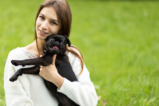 White pet owner hugs adorable pug-dog of breed 'Petit Brabancon Brussels Griffon' by hands outside in green park. Adoption of short-muzzled, human&ndash;canine bonding. Animal care. Copy space, horizontal