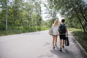 back view of tourists with backpacks and acoustic guitar walking on road near forest.