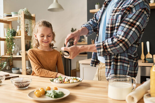 Close-up of young dad using pepper shaker to add spices in vegetable salad, he preparing together with his son in kitchen