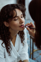 Hand of female makeup artist applying lipstick or gloss on lips of pretty bride with dark long wavy hair on wedding morning