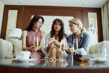 Young bride to be looking at pearl bracelet on her wrist between two friends sitting on white leather couch in living room