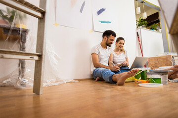 Young couple sitting on the floor choosing color via laptop for painting the wall in their home.