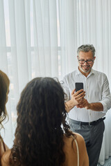 Happy mature man with smartphone taking photo of his daughter and wife standing in front of him and posing on wedding morning
