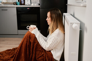 Worried sad woman sits under blanket near heating radiator with cup of tea, Rising costs in private...