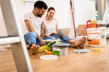 Young couple sitting on the floor choosing color via laptop for painting the wall in their home.