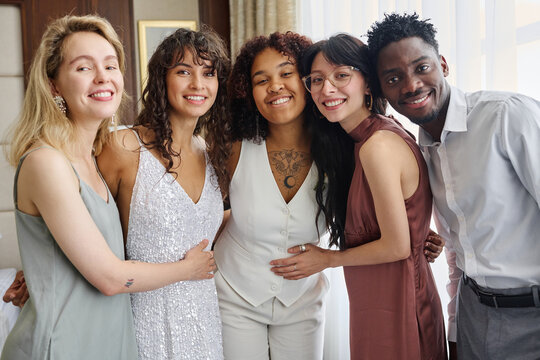 Intercultural Group Of Happy Elegant Wedding Guests Surrounding Affectionate Newlyweds In White Attire While Enjoying Home Party