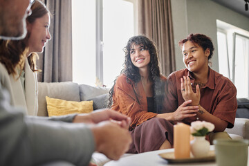Happy young multi-ethnic woman showing engagement ring on her finger while talking to parents of her girlfriend in living room