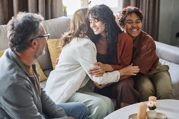 Happy young brunette woman giving hug to mother while sitting on couch next to her cheerful African American girlfriend