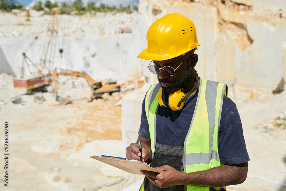 Poster young engineer in workwear making notes in working document in clipboard while writing down plan at 