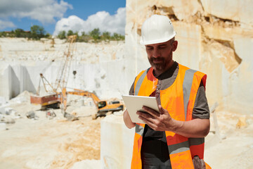 Middle aged foreman with tablet looking through online instruction at workplace while standing against wall built up of marble blocks