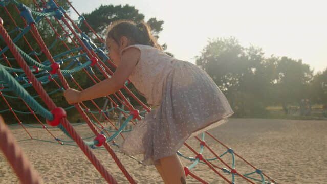 Child climbs a climbing frame on a sunny day in slow motion. Cheerful girl preschooler plays on the playground. little girl climbs up a web of ropes, a little climber.