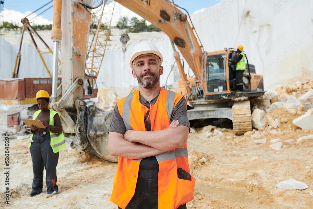Poster confident foreman in reflective vest and safety helmet looking at camera while standing on construct