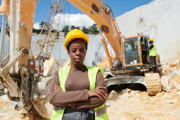 Young confident black woman in workwear and protective helmet standing with her arms crossed on...