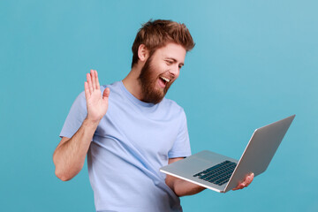 Portrait of friendly bearded man greeting a worker through a webcam and showing palm of the hand gesture with toothy smile, having online conversation. Indoor studio shot isolated on blue background.