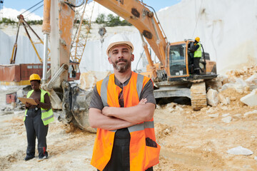 Confident foreman in reflective vest and safety helmet looking at camera while standing on...