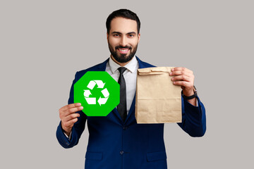 Portrait of smiling bearded man holding green recycling sign in hand and paper package, thinking...