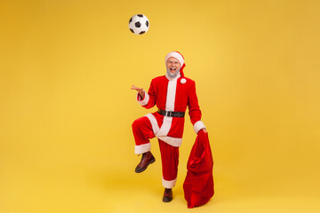Fototapeta na wymiar Full length of elderly man with gray beard wearing santa claus costume playing with soccer ball, holding red bag bags with new year presents. Indoor studio shot isolated on yellow background.