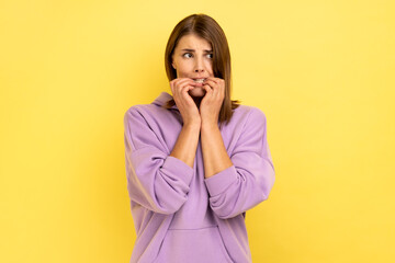 Troubles and worries. Portrait of nervous woman biting nails, terrified about problems, suffering phobia, anxiety disorder, wearing purple hoodie. Indoor studio shot isolated on yellow background.