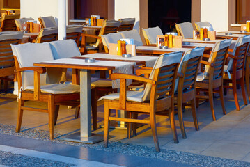 Wooden tables and chairs are neatly arranged with light tablecloths in the morning sunlight. Street cafe waiting for visitors.
