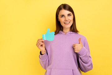 Smiling woman showing thumbs up blue paper sign, liking and recommending posts in social networks, asking to rate, wearing purple hoodie. Indoor studio shot isolated on yellow background.