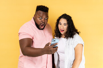 Amazed young couple in casual clothing standing together and reading unexpected message with open mouth, using mobile phone, checking social networks. Indoor studio shot isolated on yellow background.