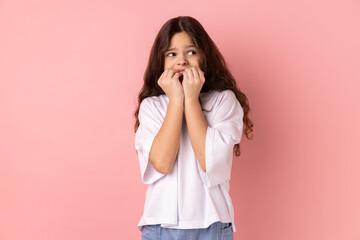 Troubles and worries. Portrait of little girl wearing white T-shirt biting nails, terrified about problems, suffering phobia, anxiety disorder. Indoor studio shot isolated on pink background.