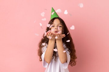 Portrait of little girl wearing white T-shirt and party hat blowing heart shape glitter, enjoying...