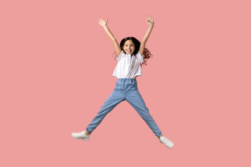 Fototapeta na wymiar Full length portrait of charming beautiful little girl wearing white T-shirt jumping in air and raised arms, expressing excitement, having fun. Indoor studio shot isolated on pink background.