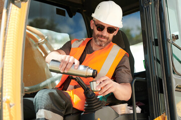 Male driver of construction machine pouring hot tea or water from thermos over gearshift while...