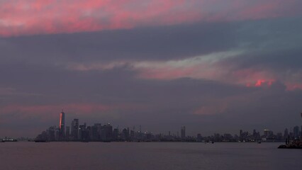 Timelapse of getting dark above the Manhattan skyscrapers in the evening seen from the American Veterans Memorial Pier. Sailing ferries. Clouds are lightened with pink and purple by the sun. USA, NYC