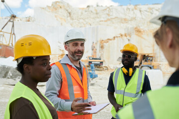 Confident foreman in workwear and hardhat giving instructions to workers or explaining them working points at meeting
