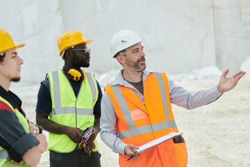 Middle aged foreman in workwear and safety helmet pointing at new industrial equipment while explaining his subordinates how to use it