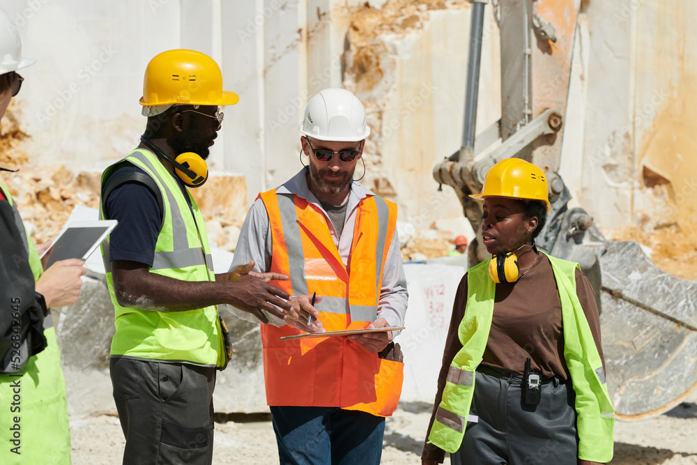 Poster Group of interracial workers in hardhats and uniform discussing working plan at meeting while confident foreman pointing at document