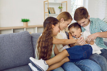 Cheerful family with children having fun together and fooling around tickling on sofa in living room. Funny young dad, mom and their daughter tickle little boy. Concept of leisure time with family. - obrazy, fototapety, plakaty