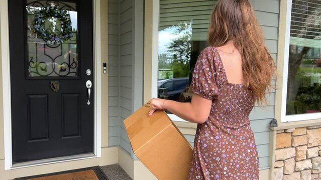 A teenager stands in a short summer dress against the backdrop of a private house in Canada, she holds a cardboard box in her hands and looks at the closed door She has long blonde curly hair.