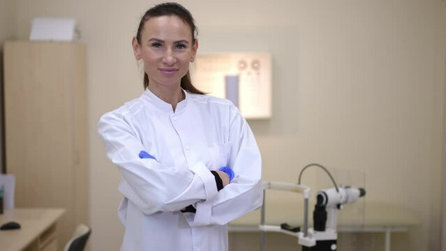 Beautiful intelligent female ophthalmologist crossing hands looking at camera standing in hospital indoors. Live camera zoom in to confident Caucasian woman posing in slow motion in medical clinic