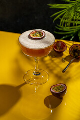 Glass of Porn Star Martini cocktail with passionfruit on yellow table with dark background, raw fruits around. Summer exotic fruit alcohol refreshing drink with foam. Copy space.