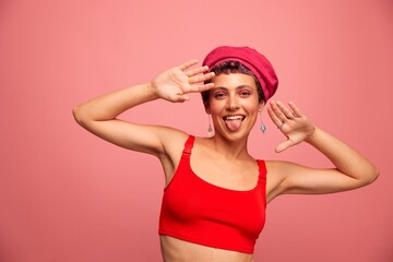 Obraz premium Young athletic woman with a short haircut and purple hair in a red top and a pink hat with an athletic figure smiles and grimaces looking at the camera on a pink background