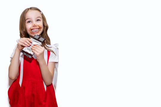 Happy little girl in a red dress eats a bar of chocolate on a white background. Funny girl painted her face with chocolate. Portrait of a child with a bar of chocolate. Free space for text