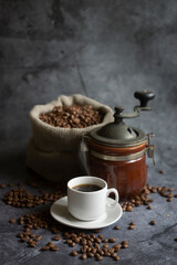 Hot coffee in a white coffee cup, a coffee grinder and a lot of coffee beans scattered around, on a dark background.